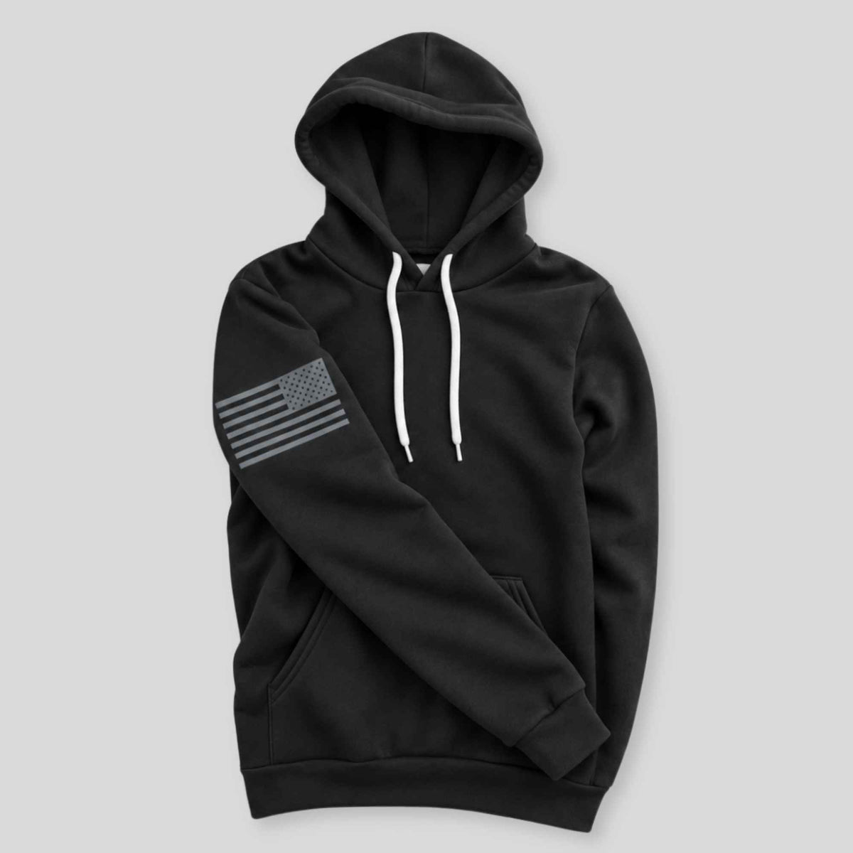 A black pullover hoodie with white drawstrings and a distressed gray American flag graphic on the left sleeve, displayed against a solid light gray background.