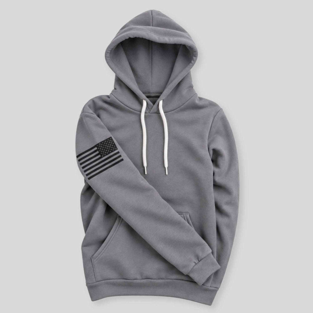 A gray pullover hoodie with white drawstrings and a black distressed American flag graphic on the left sleeve, displayed against a solid light gray background.