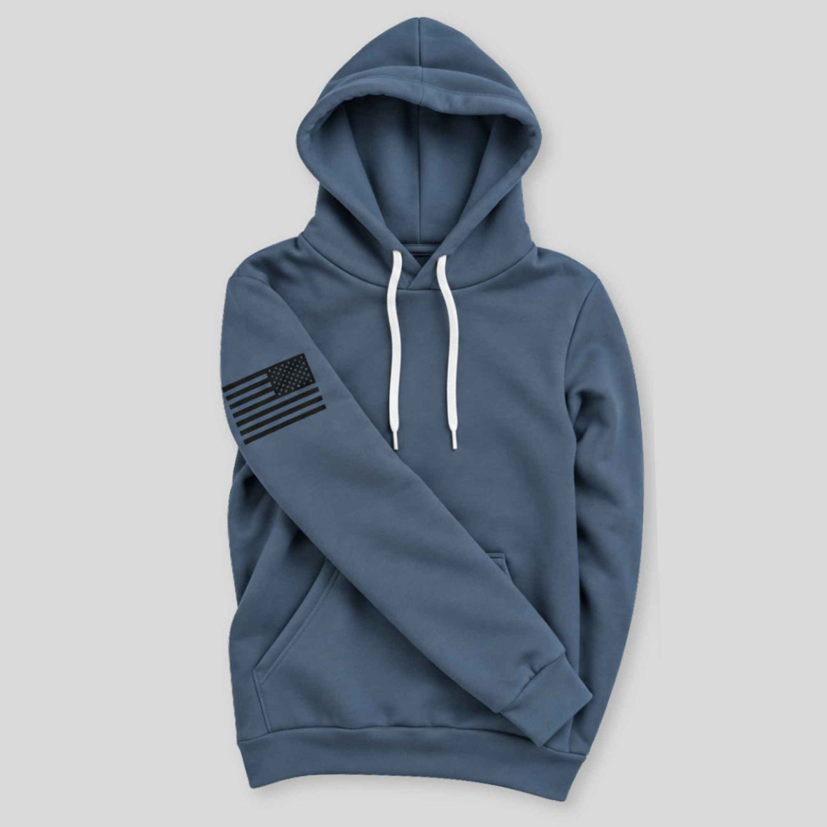 A slate blue pullover hoodie with white drawstrings and a black distressed American flag graphic on the left sleeve, displayed against a solid light gray background.