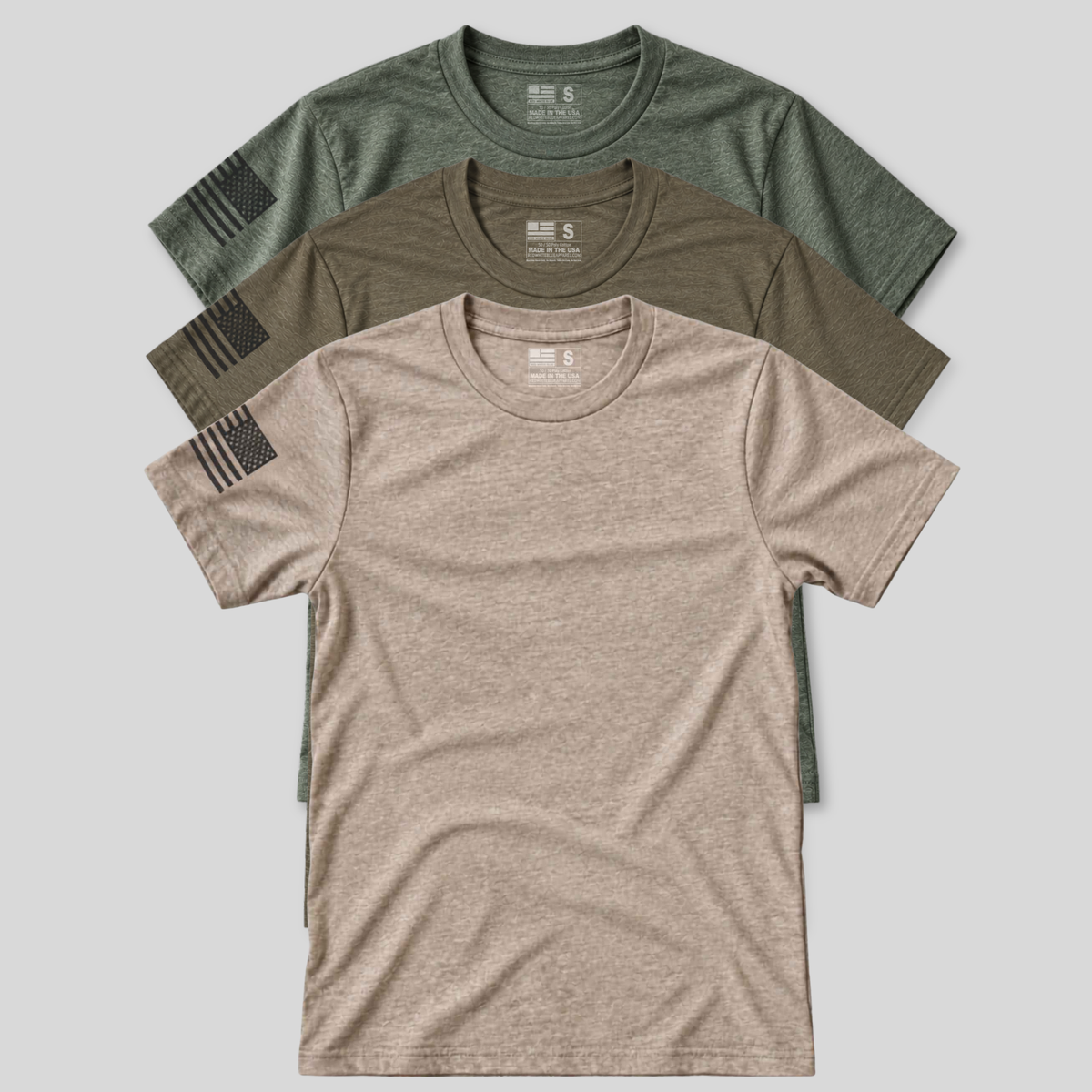 Three short-sleeve crewneck t-shirts in heathered olive green, olive brown, and tan, arranged in a stacked fan layout on a light gray background. Each shirt features a black distressed American flag graphic printed on the left sleeve.