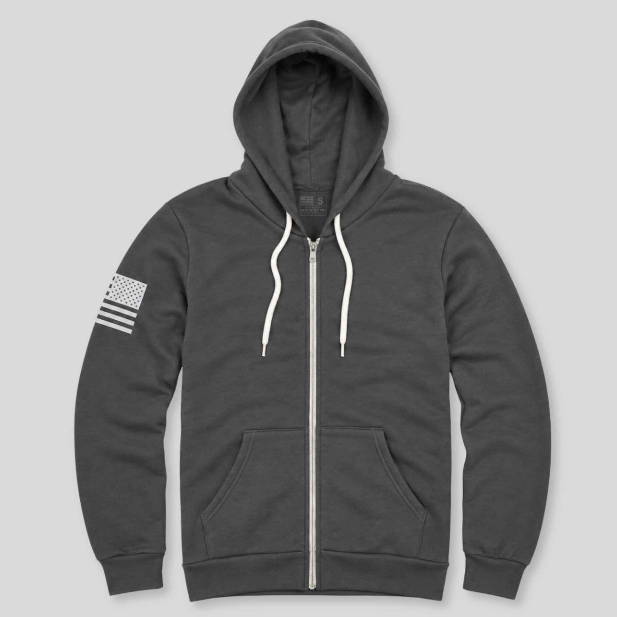 A dark gray zip-up hoodie with white drawstrings, a white front zipper, and a white American flag graphic on the left sleeve, displayed against a solid light gray background.