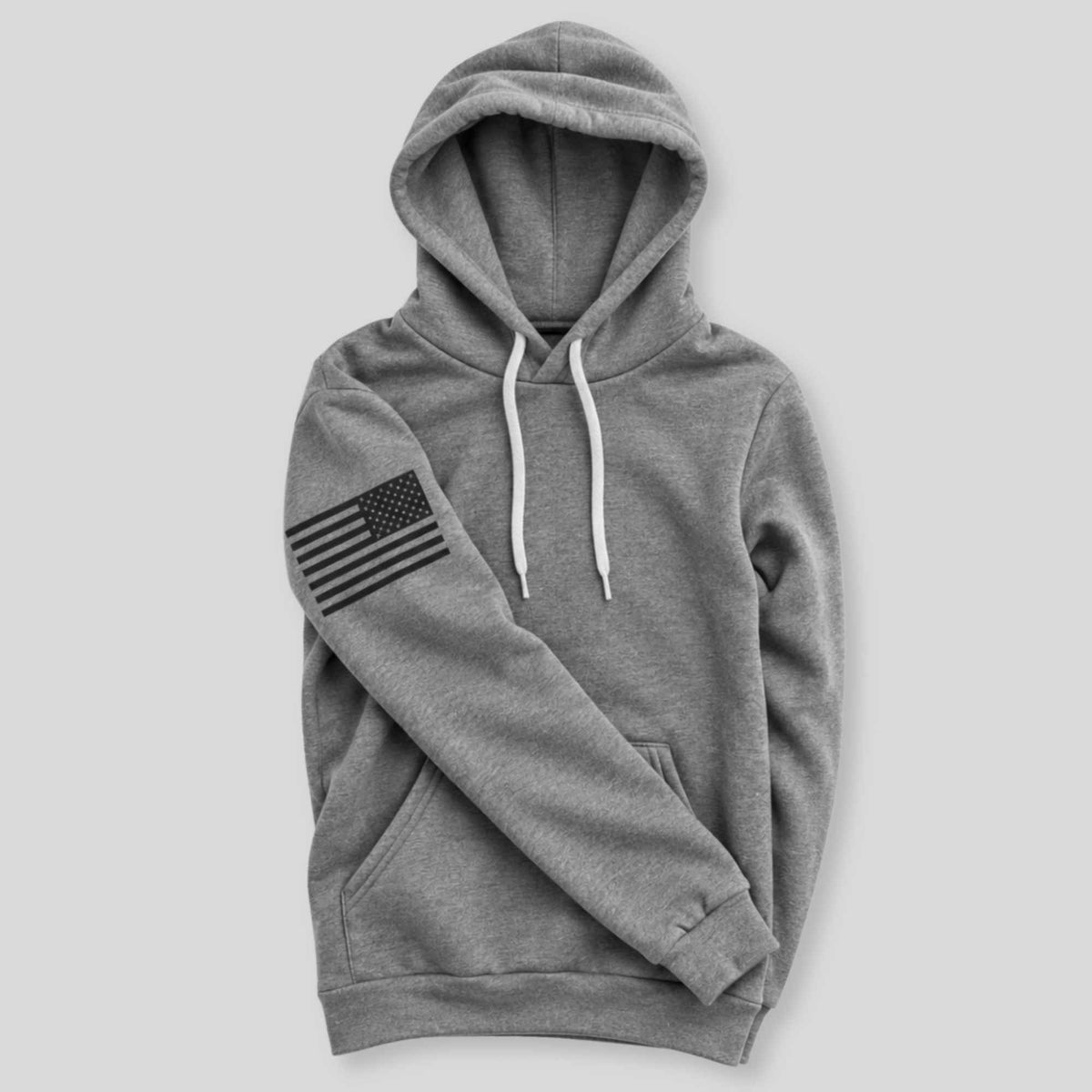 A heather gray pullover hoodie with white drawstrings and a black American flag graphic printed on the left sleeve, displayed against a solid light gray background.