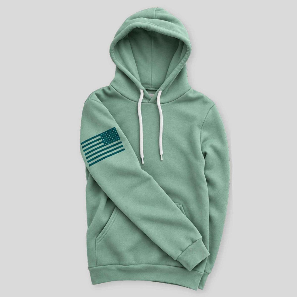 A sage green pullover hoodie with white drawstrings and a dark green American flag graphic on the left sleeve, displayed against a solid light gray background.