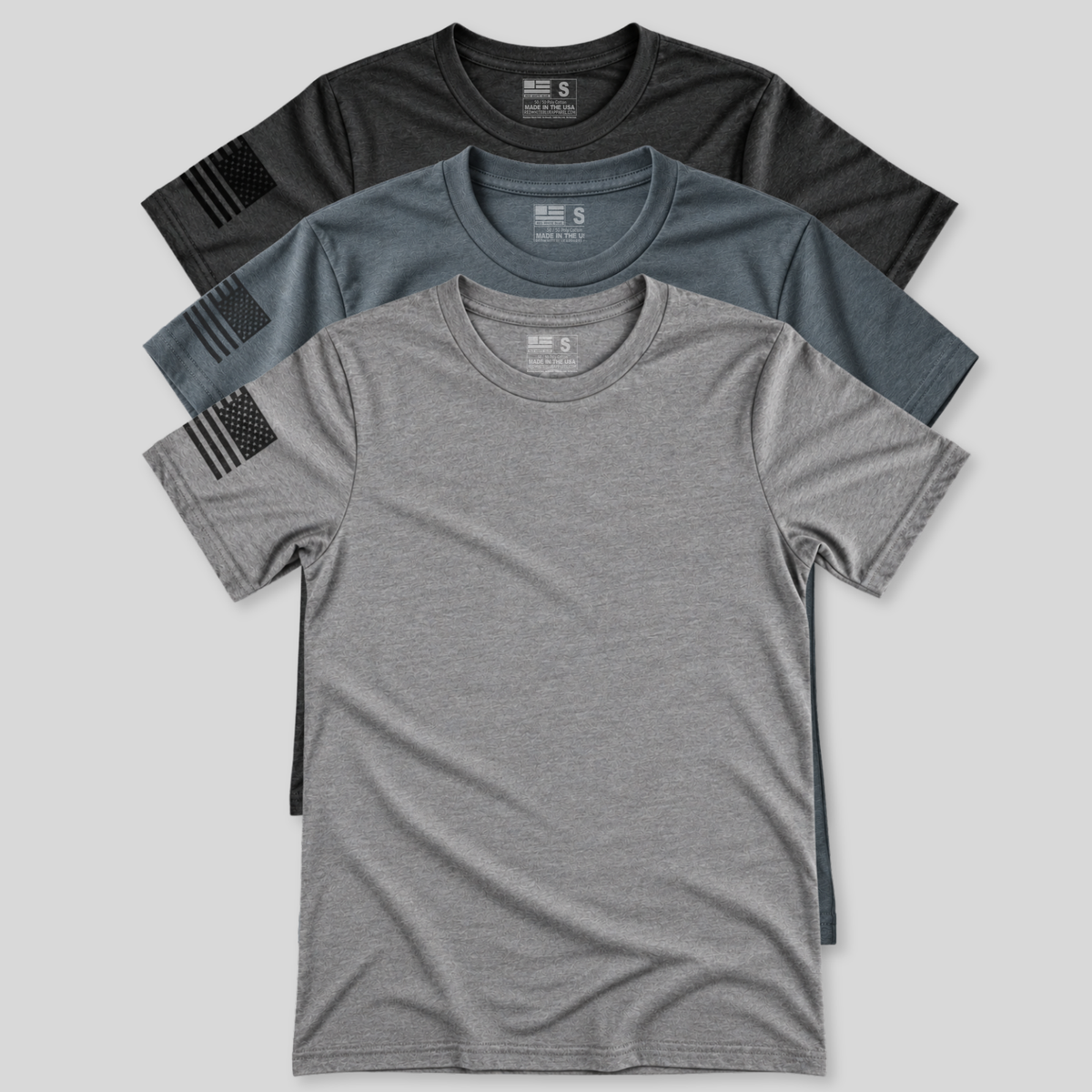 Three short-sleeve crewneck t-shirts stacked in a fan arrangement on a light gray background. The shirts are heather gray, heather blue-gray, and dark charcoal/black, each featuring a black distressed American flag patch on the right sleeve.